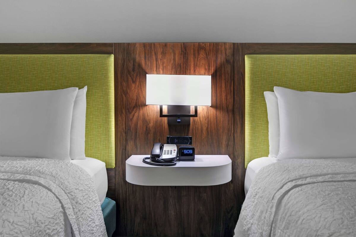 A hotel room with two beds, a nightstand with a phone and clock, and a wall-mounted lamp.