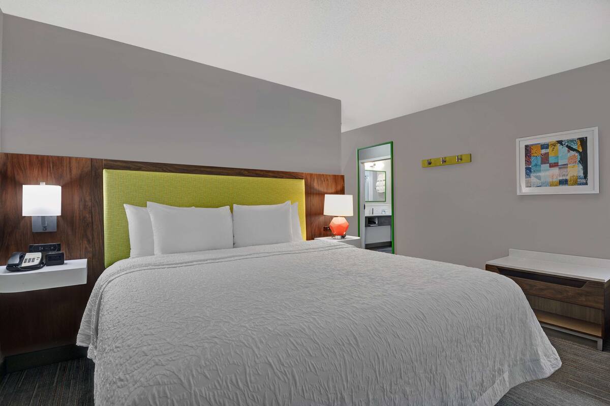 A hotel room with a large bed, a bedside table with a lamp, a telephone, a mirror, and a framed artwork on the wall.