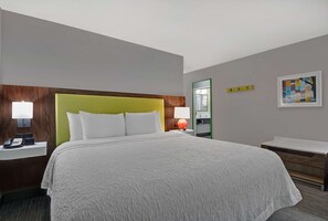 A hotel room with a large bed, a bedside table with a lamp, a telephone, a mirror, and a framed artwork on the wall.
