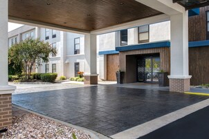 Un espacio al aire libre techado con un sendero pavimentado, la entrada de un edificio con una puerta de madera y un pequeño árbol.