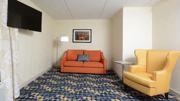 A hotel room with a sofa, armchair, patterned carpet, a flat-screen TV, a framed picture, and a floor lamp.