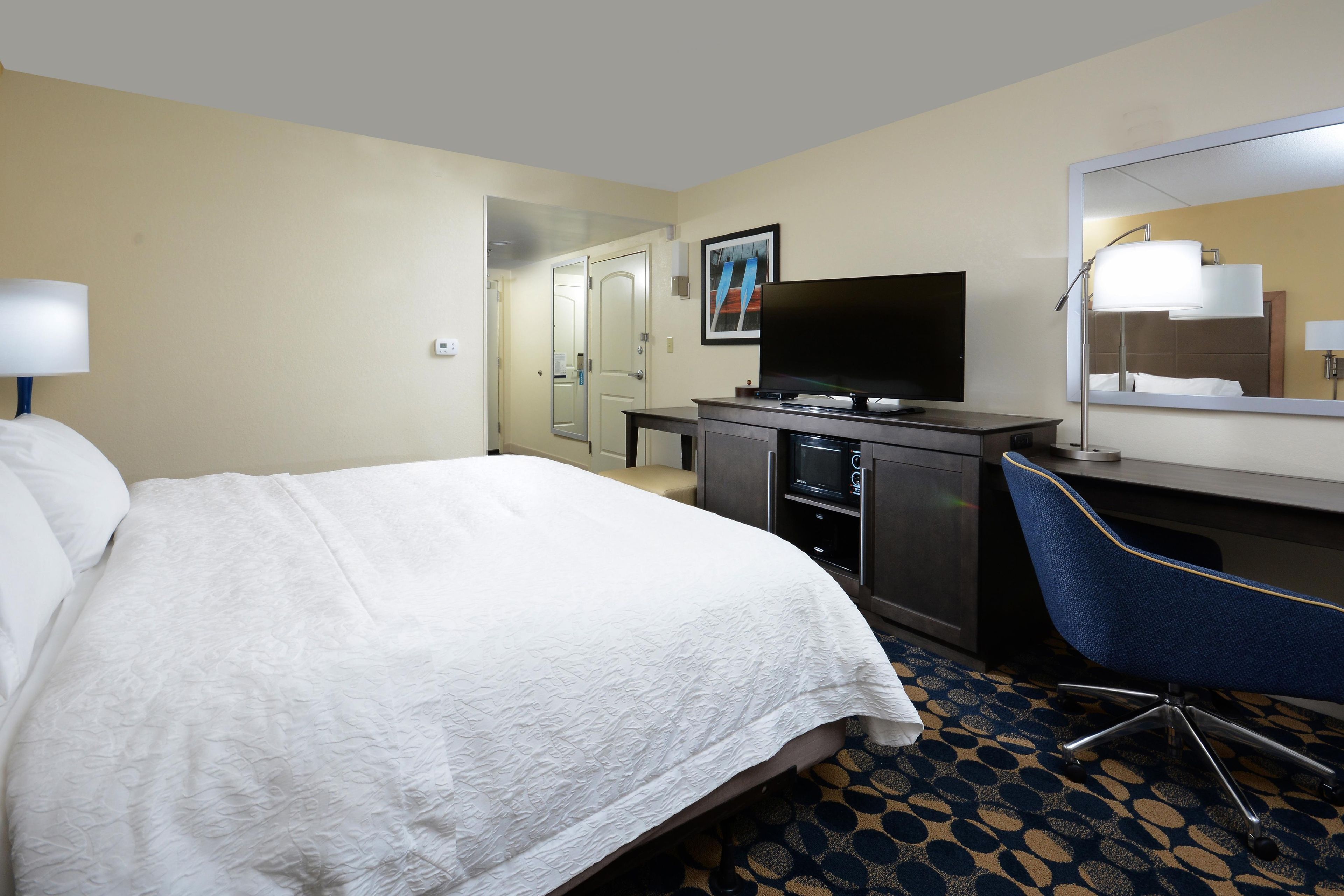 Une chambre d’hôtel avec un lit, un bureau, une télévision et un miroir dans la salle de bain.