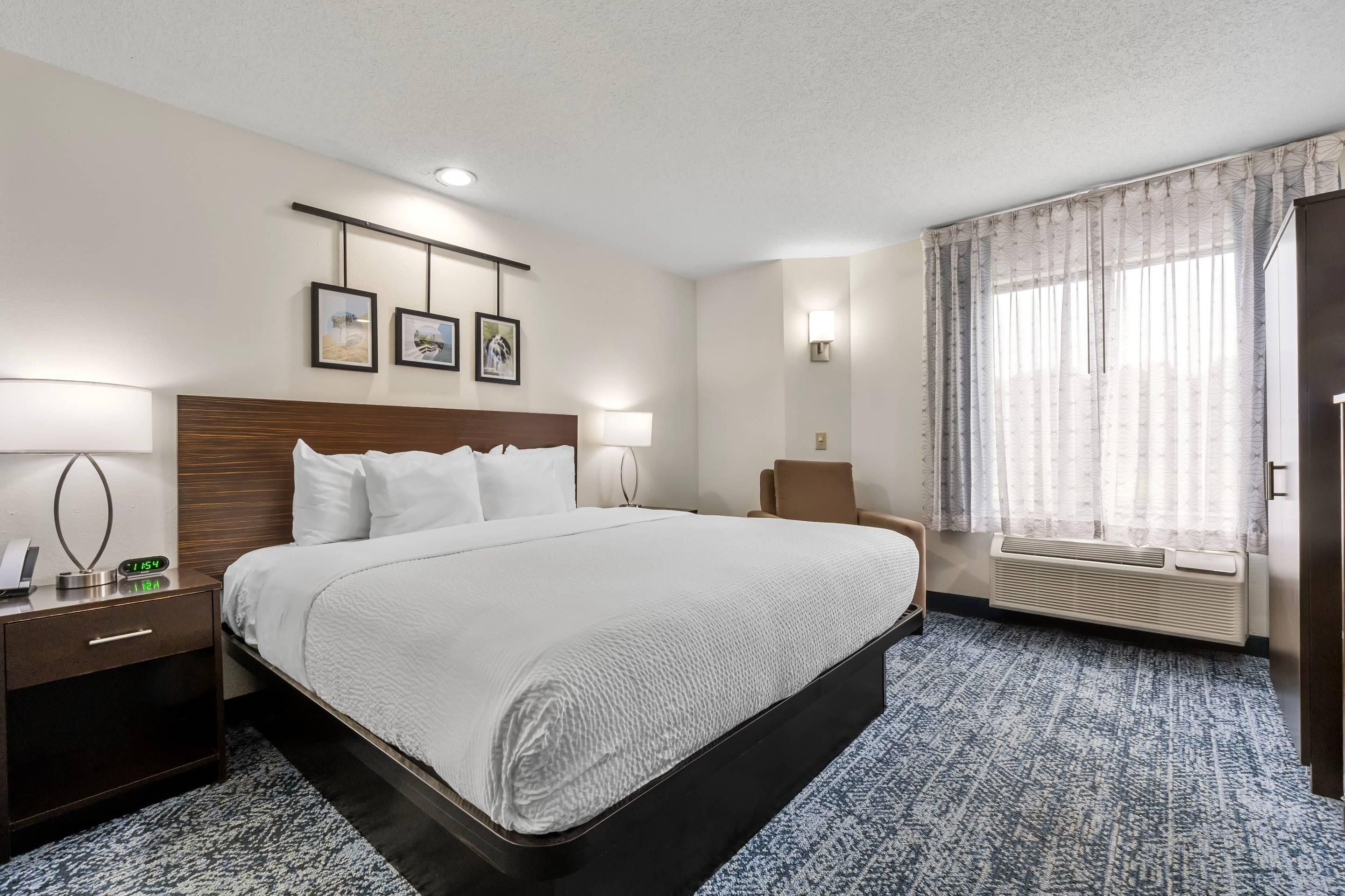 Una habitación de hotel con una cama grande, mesitas de noche, un escritorio, una silla, una ventana con cortinas y cuadros en la pared.