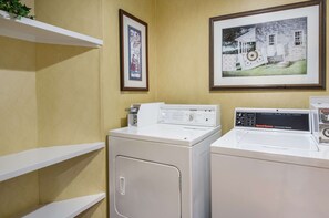 A laundry room with two front-loading washers and dryers, white shelves, and framed pictures on the wall. - Sleep Inn Hanes Mall (Winston-Salem)