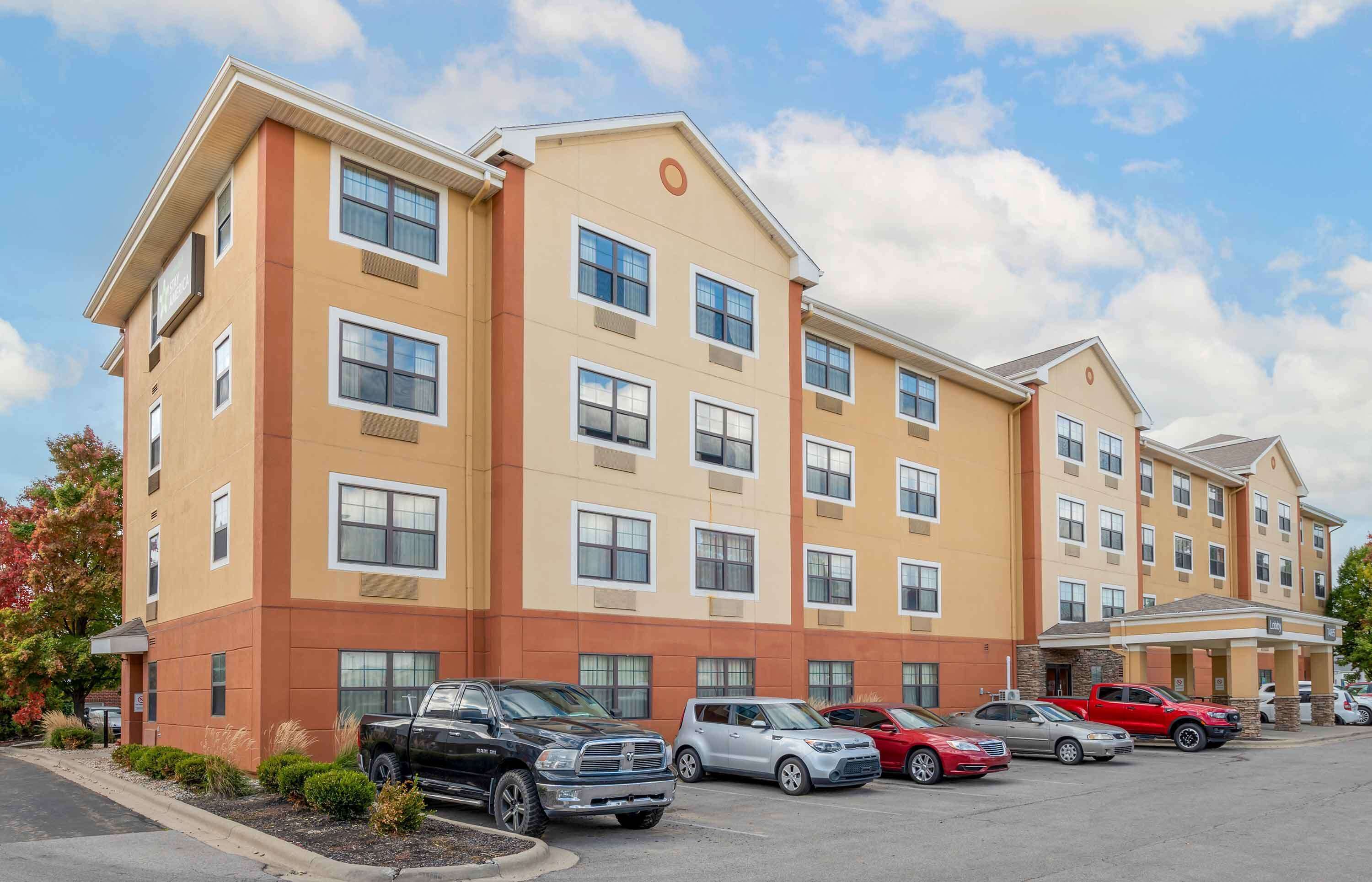 Un hôtel à plusieurs étages, doté d’une façade beige et marron, de nombreuses fenêtres et d’un parking où sont stationnés divers véhicules.