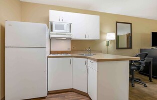 Una cocina compacta con armarios blancos, microondas, refrigerador, pileta y un pequeño escritorio con silla.