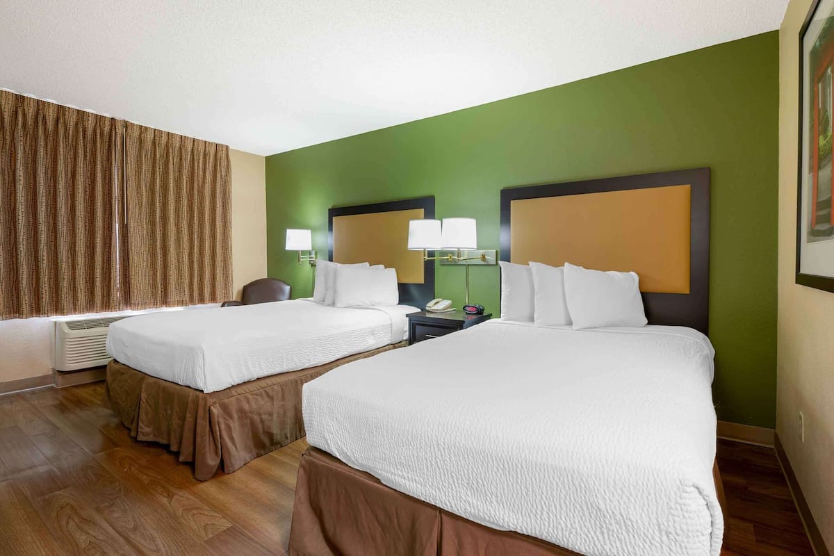 A hotel room with two beds, a large window with curtains, a desk with a lamp, and a framed picture on the wall.
