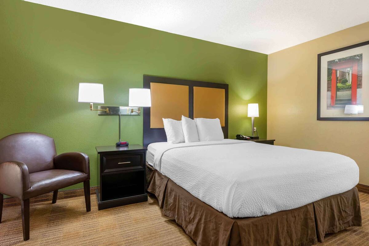A hotel room with a bed, a chair, a nightstand, and a lamp.