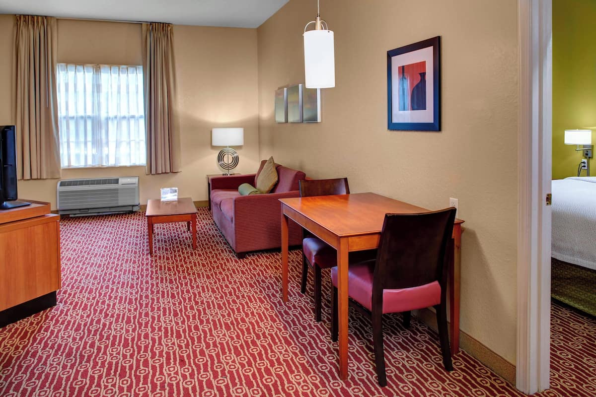 A hotel room with a red patterned carpet, a wooden desk, a red sofa, a television, and a bed.