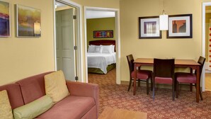 A hotel room with a red sofa, a wooden dining table with four chairs, a bed with white bedding, and a patterned carpet. - Towneplace Suites By Marriott Cleveland Westlake (Westlake)