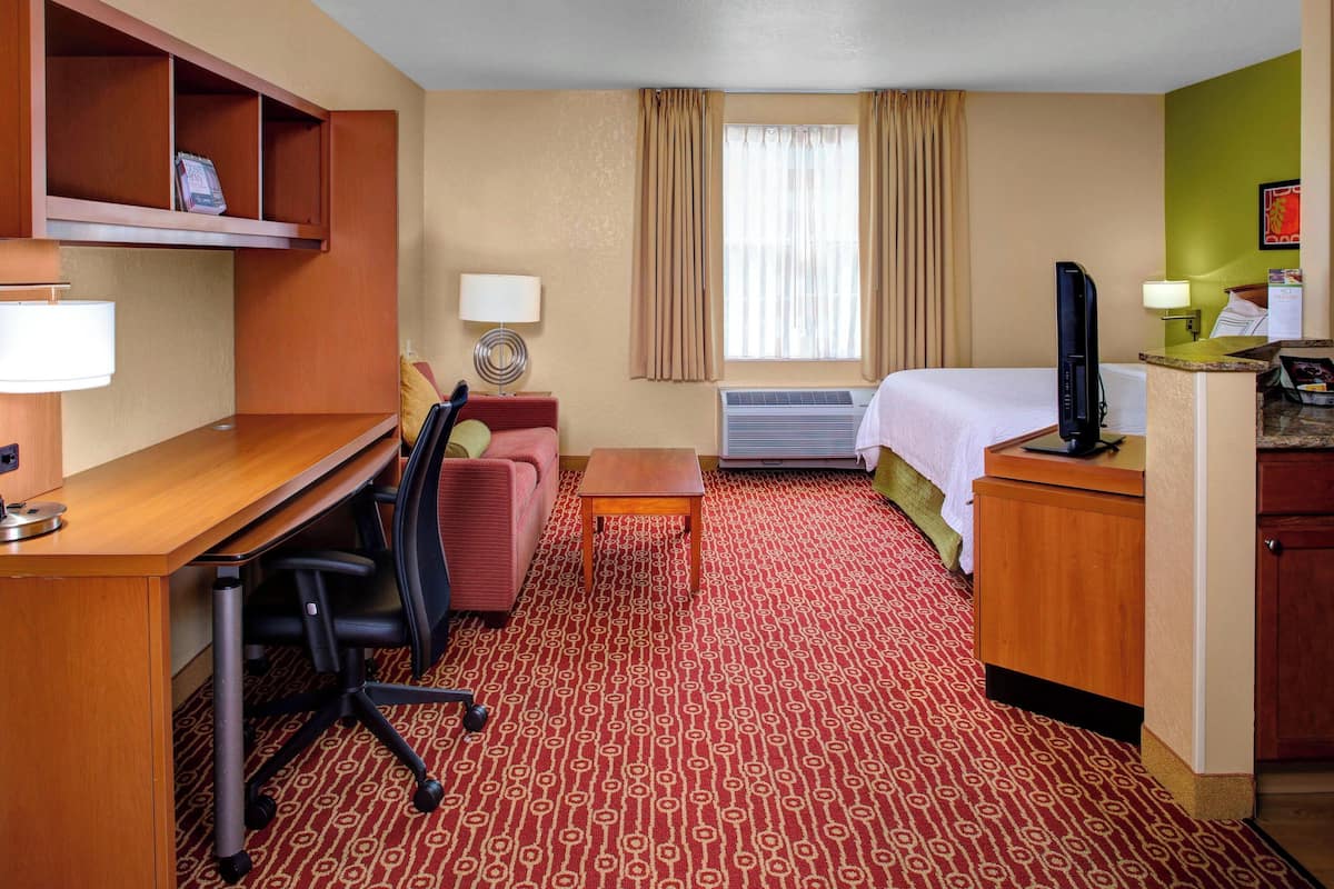 A hotel room with a desk, a chair, a bed, a TV, a lamp, and a small table.