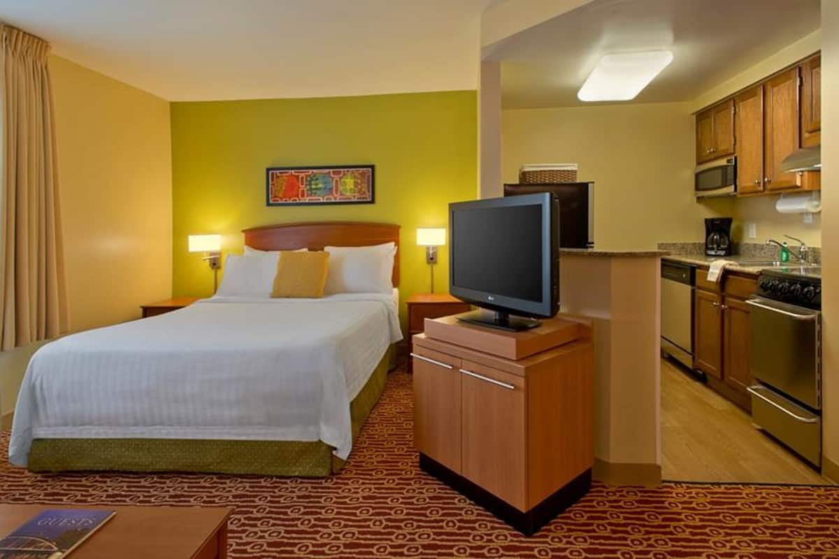 A hotel room with a bed, a television, a kitchenette, and a patterned carpet.