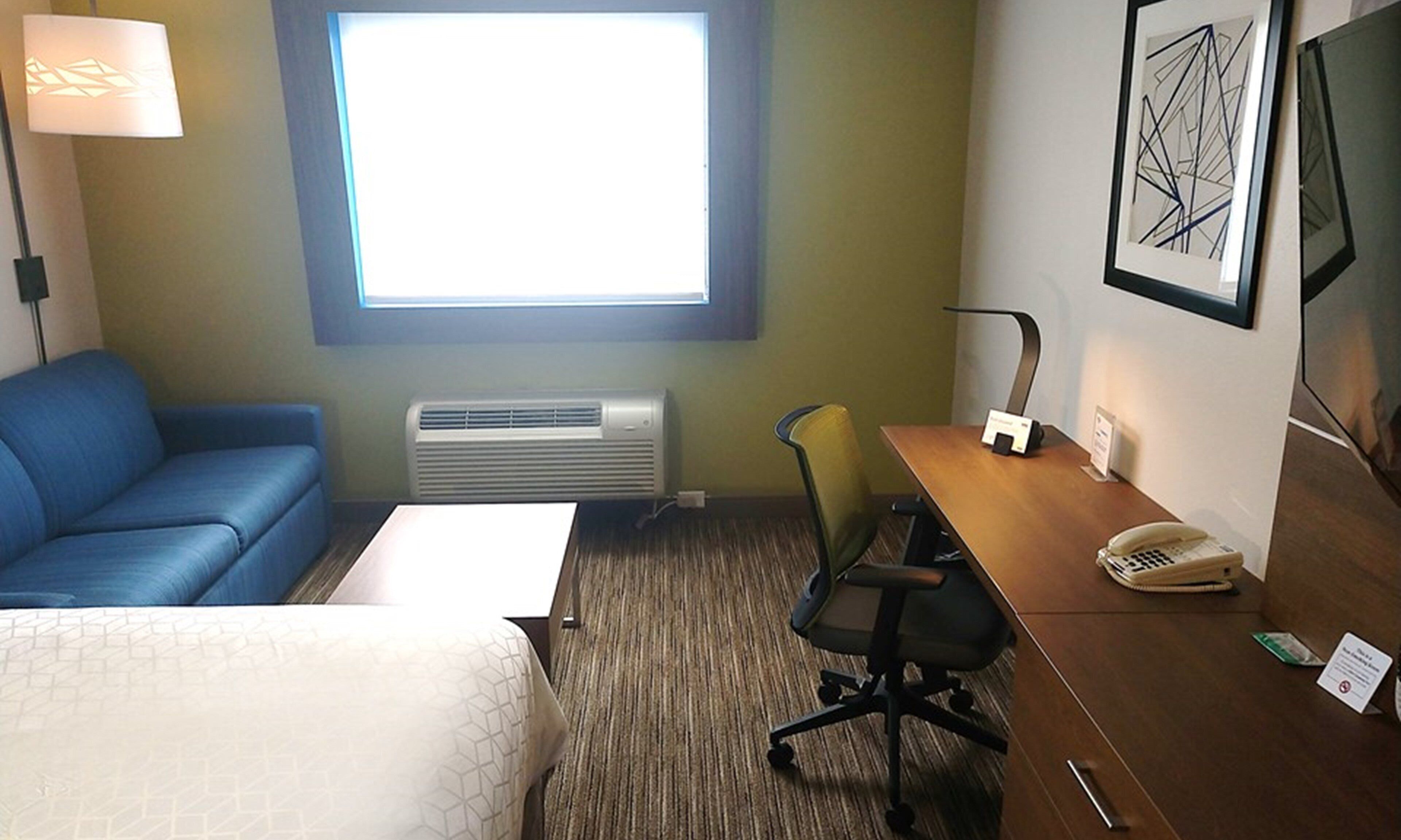 Habitación de hotel con cama, escritorio con silla, teléfono, aire acondicionado de pared y ventana con persianas, además de una obra de arte abstracta enmarcada.