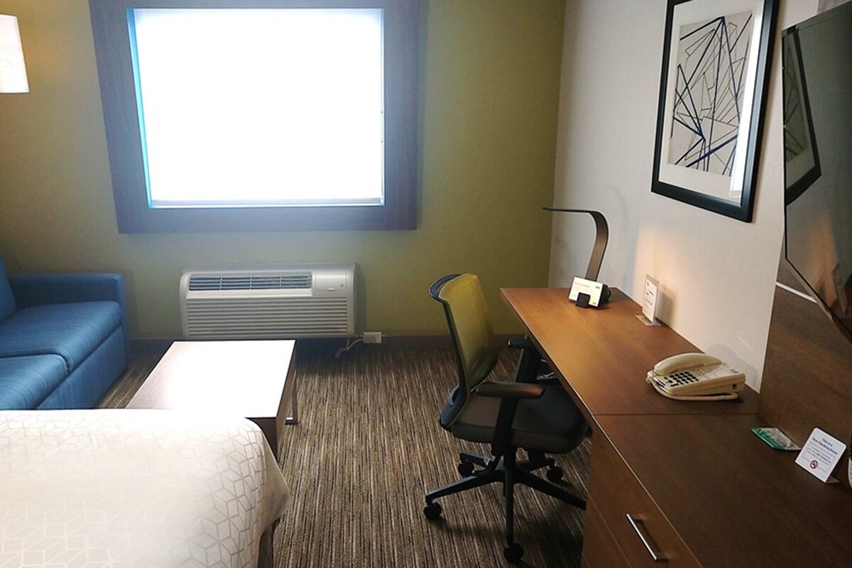 Ein Hotelzimmer mit einem Bett, einem blauen Sofa, einem Schreibtisch mit Stuhl, einem Telefon und einem Fenster mit Jalousien.