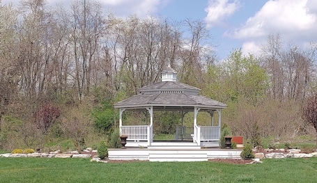 A gazebo with a white roof and white railings, surrounded by a well-maintained lawn and a dense forest in the background.