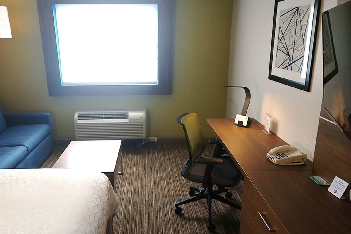 Ein Hotelzimmer mit einem Bett, einem blauen Sofa, einem Schreibtisch mit Stuhl, einem Telefon und einem Fenster mit Jalousien.