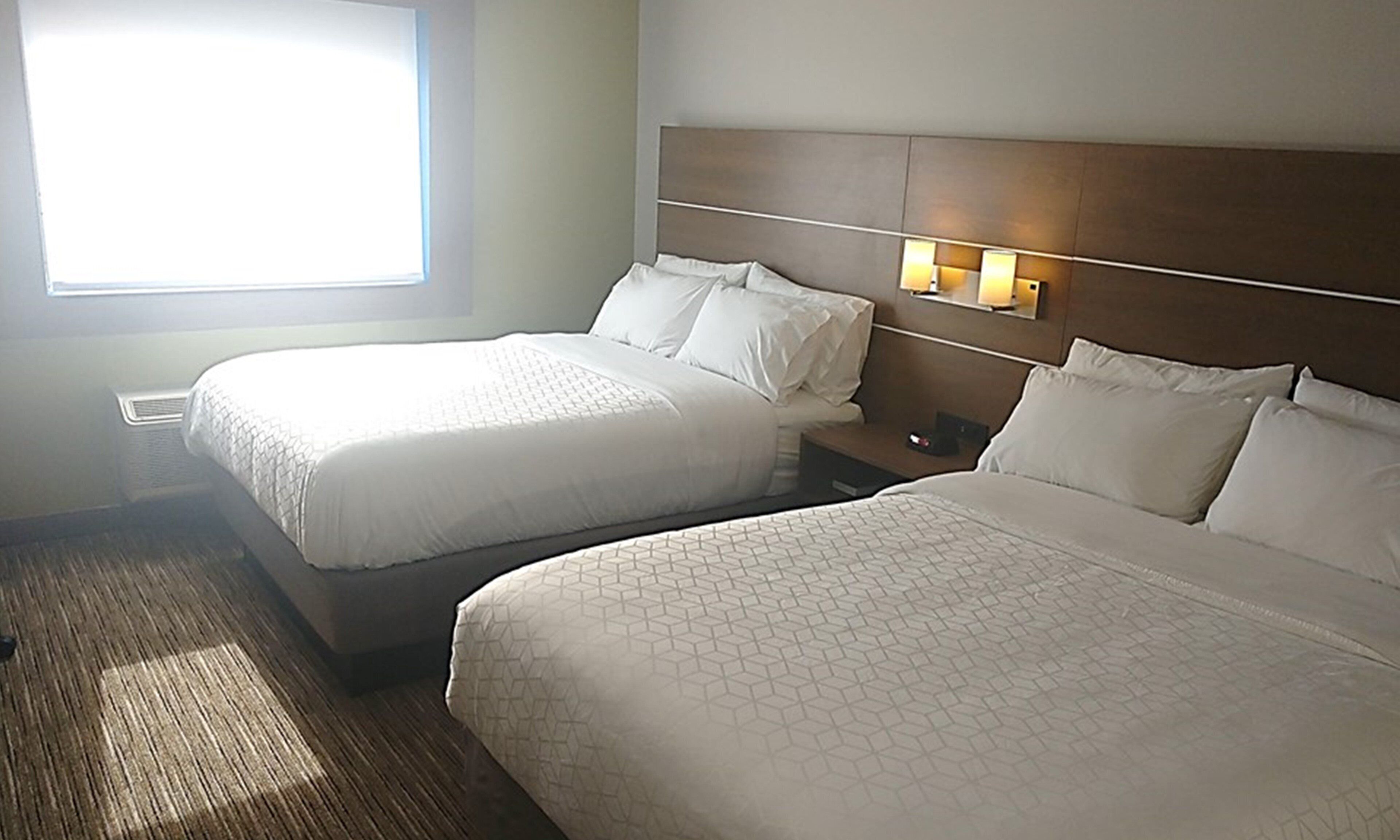 A hotel room with two beds, a nightstand, a window with blinds, and a wall-mounted light fixture.