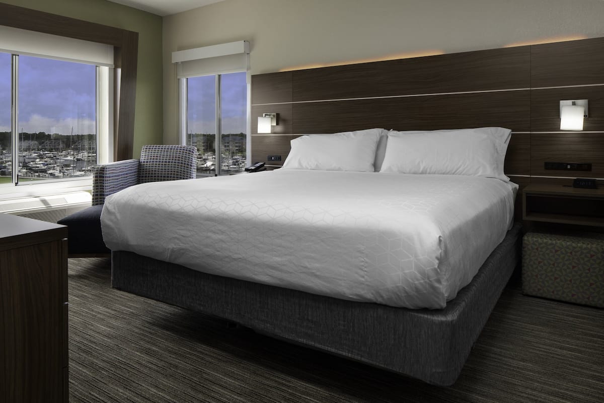 A modern hotel room with a large bed, a desk, and a chair. There are two bedside tables with lamps and a large window offering a city view.