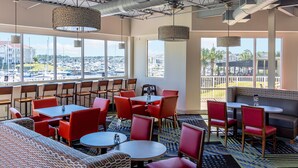A spacious indoor area with multiple tables and chairs, large windows offering views of a marina and buildings, and a high ceiling with visible ductwork. - Holiday Inn Express N.Myrtle Beach- Little River by IHG (Little River)