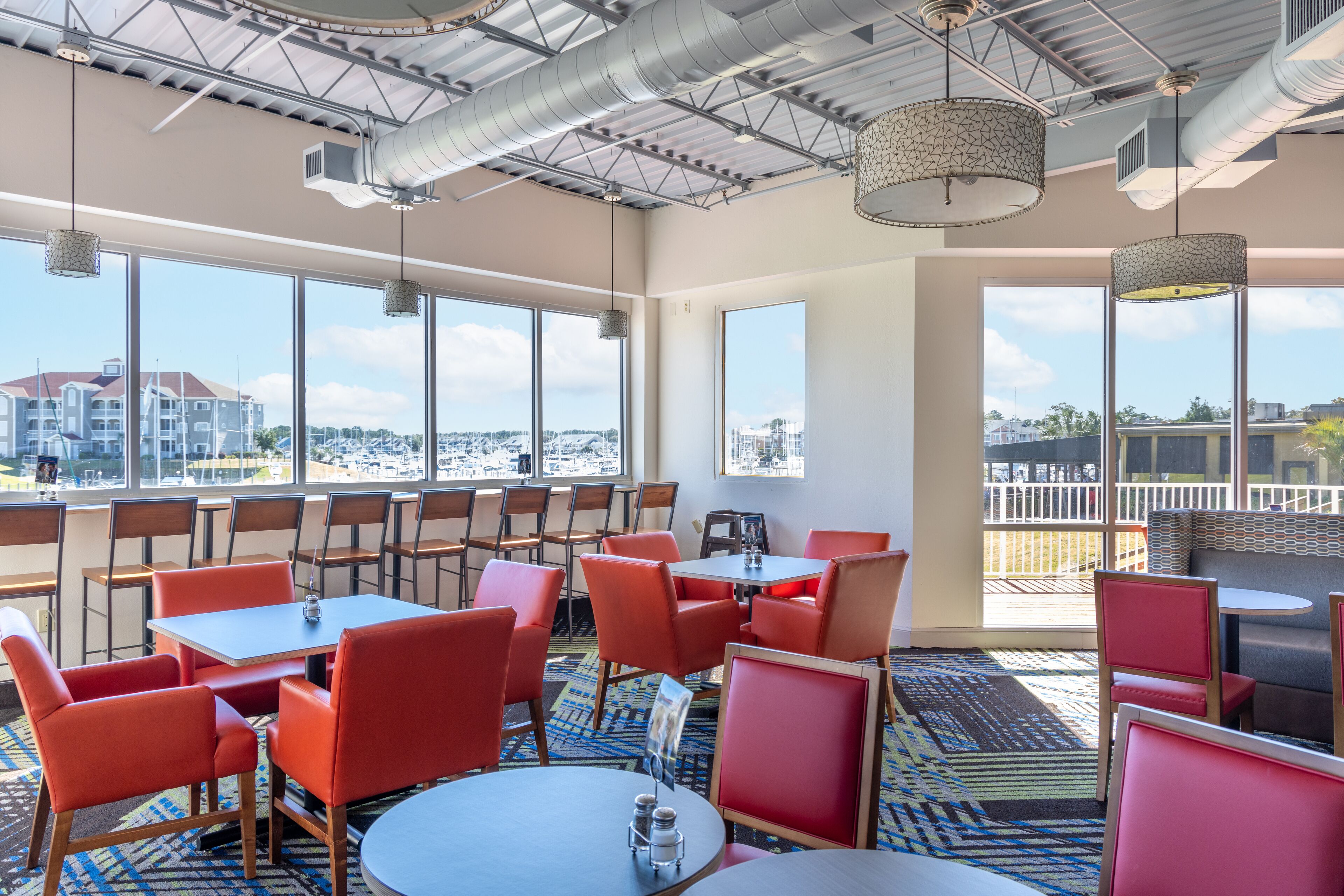 A modern dining area with red chairs and tables, large windows with a city view, and a patterned carpet.