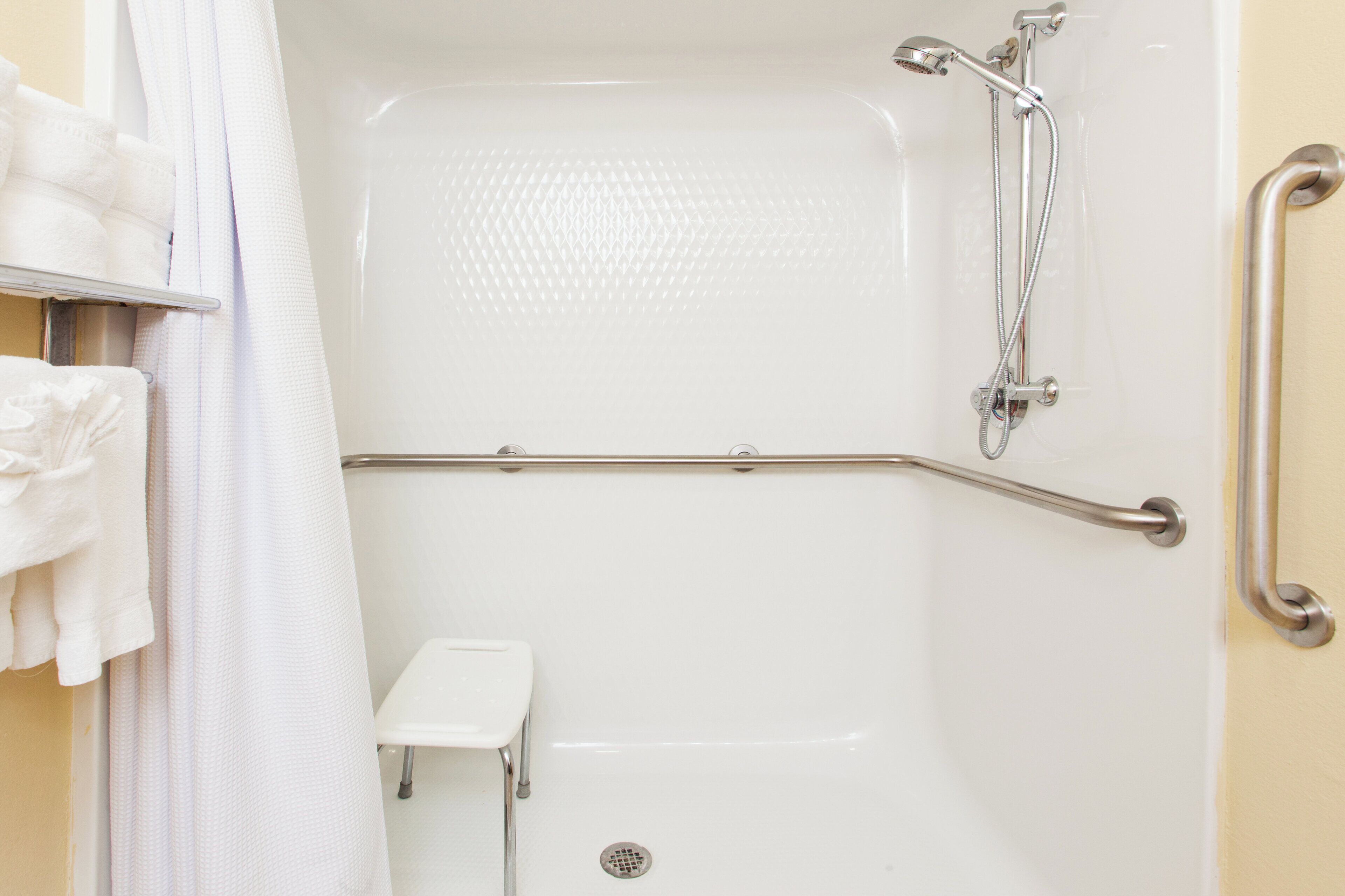 Baño con ducha a nivel, barras de apoyo y un pequeño banco blanco.