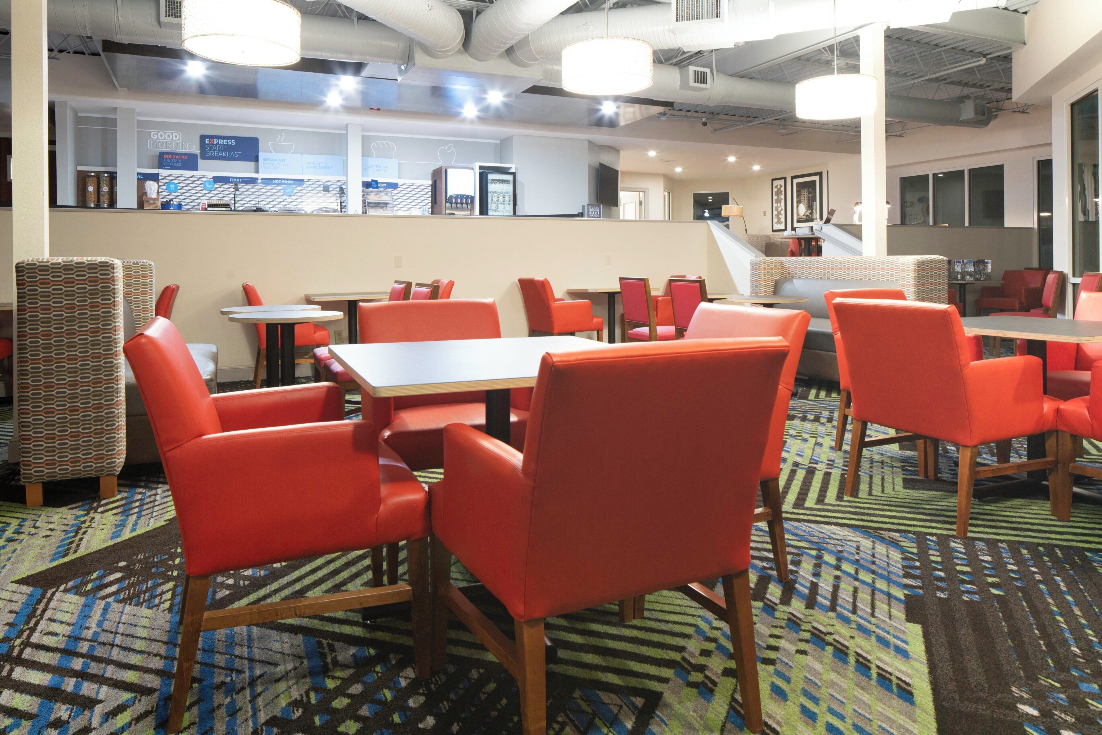 A modern dining area with red chairs and tables, a patterned carpet, and a clean, well-lit space with a visible kitchen area in the background.