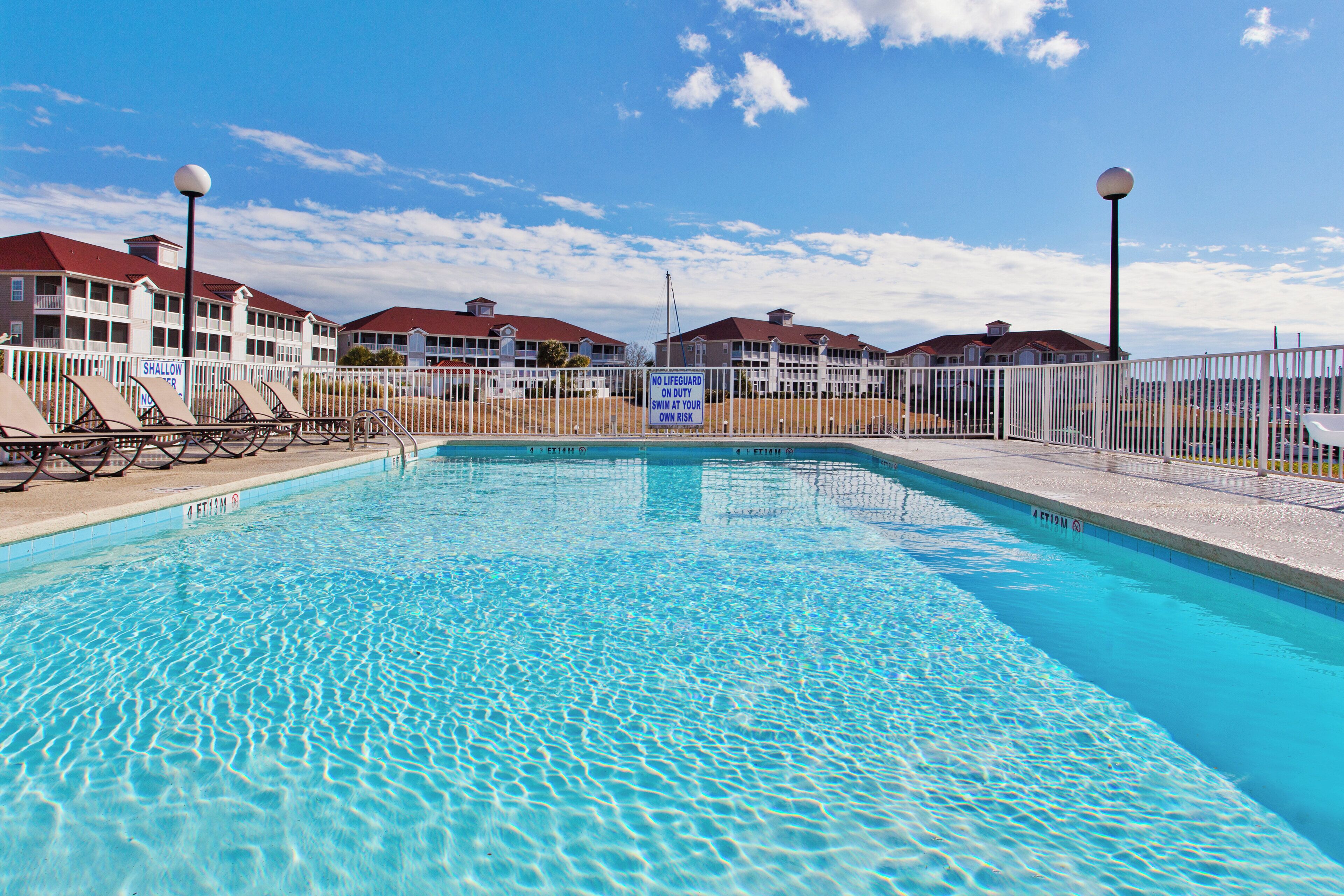 A swimming pool with a clear blue water surface, surrounded by a white fence, and a building with multiple balconies in the background.