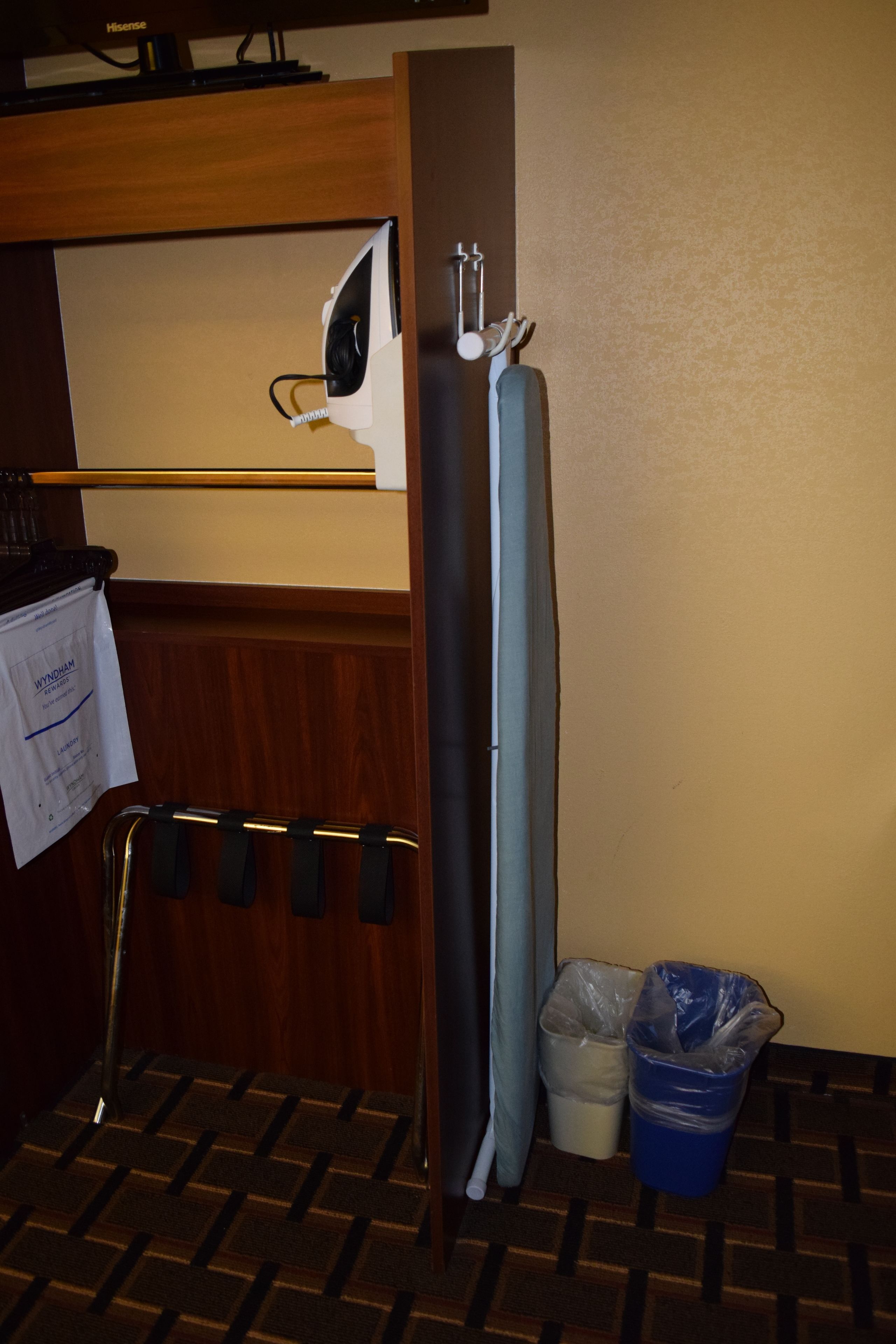 Habitación de hotel con piso de madera, tabla de planchar sujeta a la pared, un pequeño cesto de basura y un perchero para toallas.