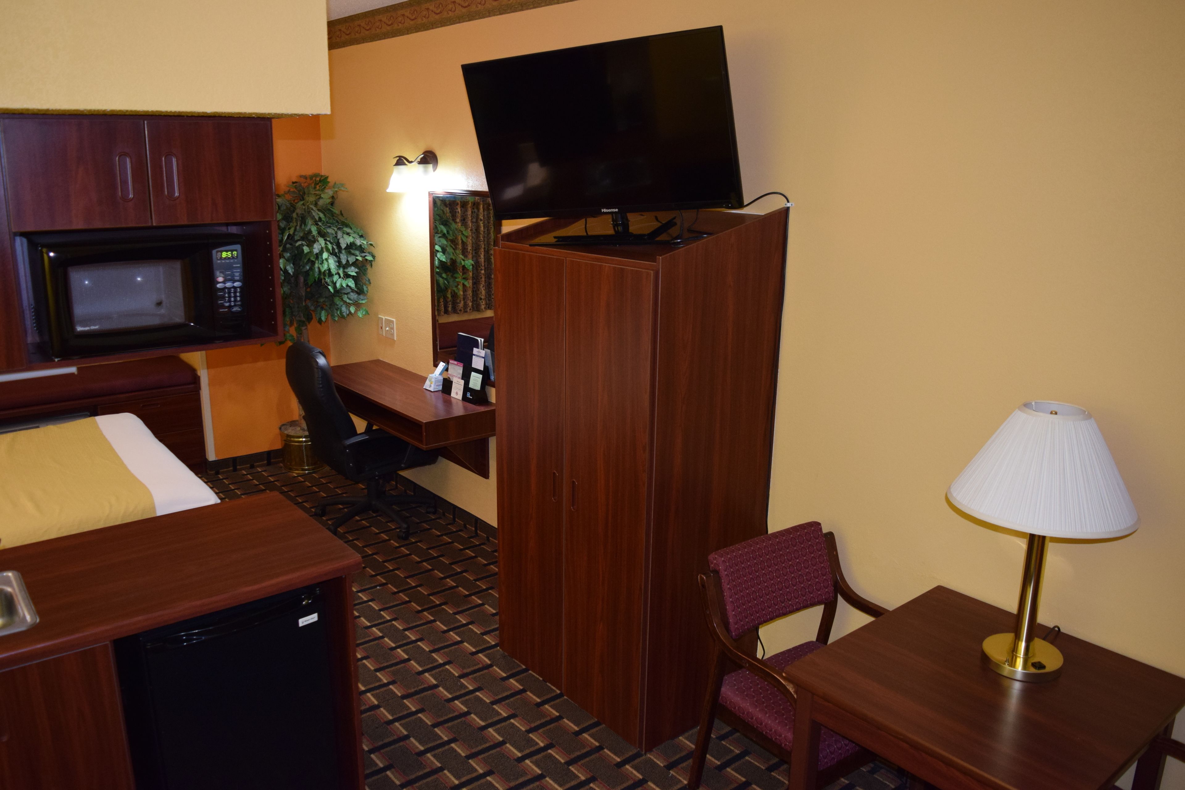 Una habitación de hotel con una cama, un escritorio con computadora, un televisor sobre un mueble, una lámpara y un microondas.
