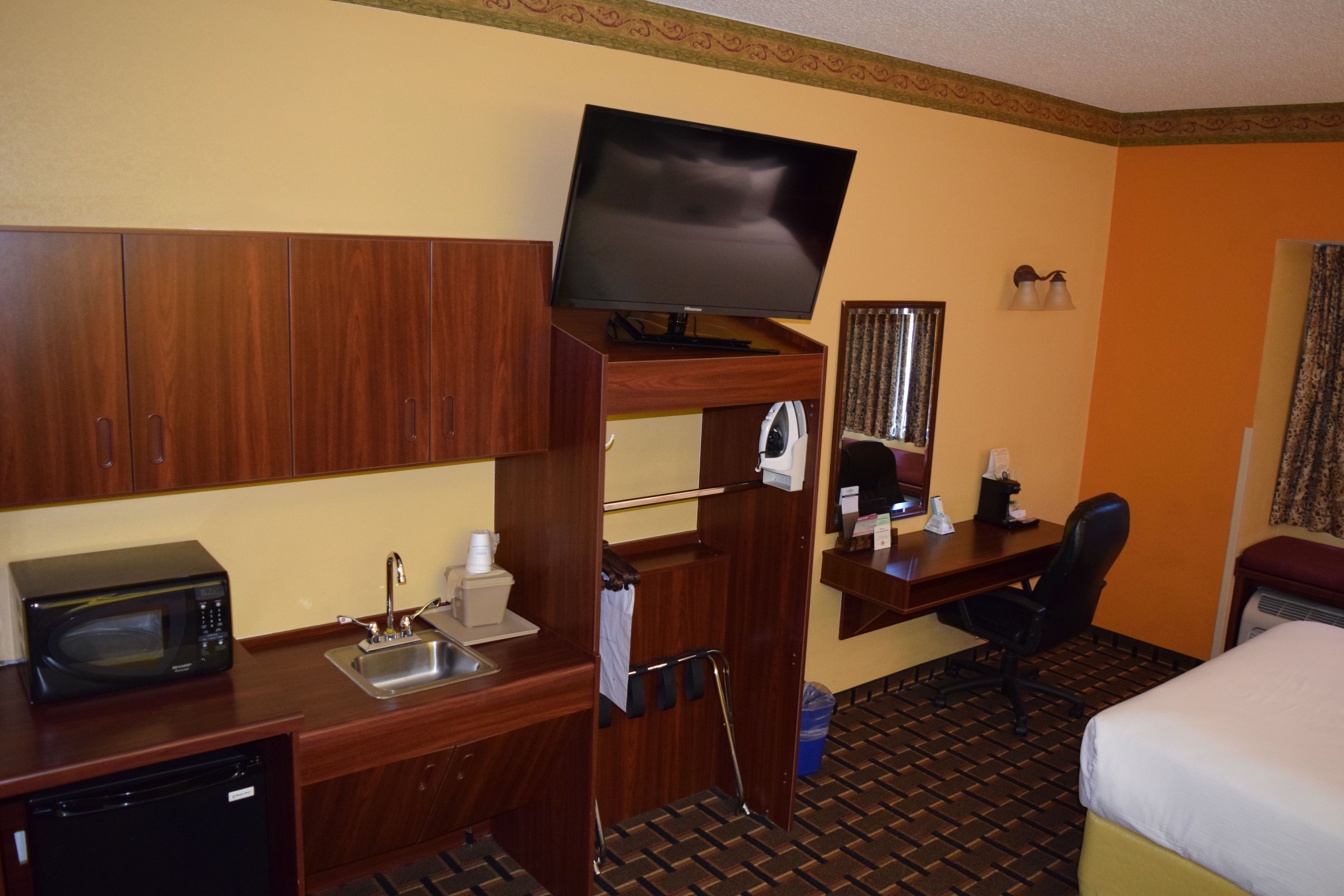 Habitación de hotel con un escritorio de madera, un televisor en un soporte de madera, microondas, lavamanos, una silla y una cama.