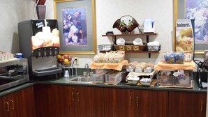 Un espace de petit-déjeuner en libre-service avec un distributeur de jus de fruits, divers produits alimentaires et un panneau indiquant la présence d'une boulangerie.