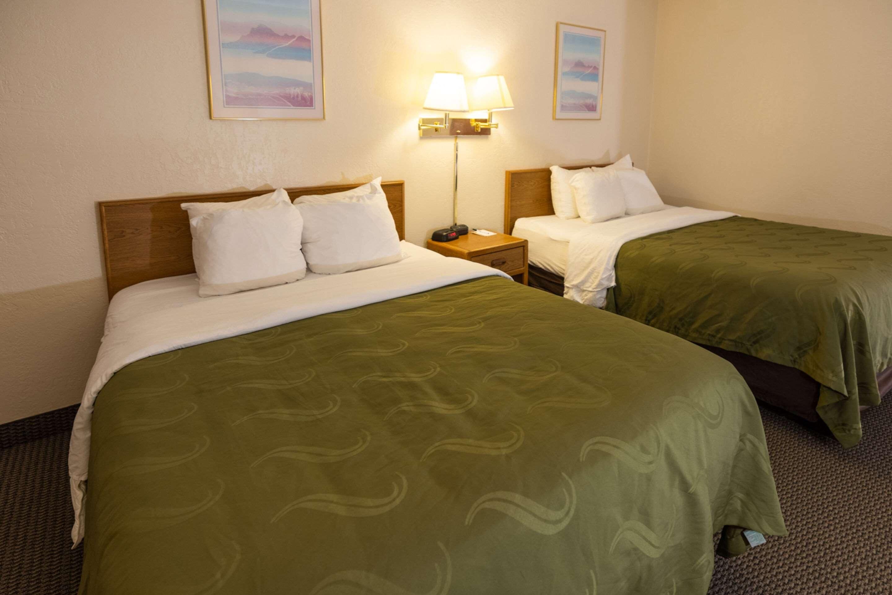 A hotel room with two beds, each with a green bedspread, white pillows, and a wooden headboard. There are two wall-mounted lamps on either side of the beds.