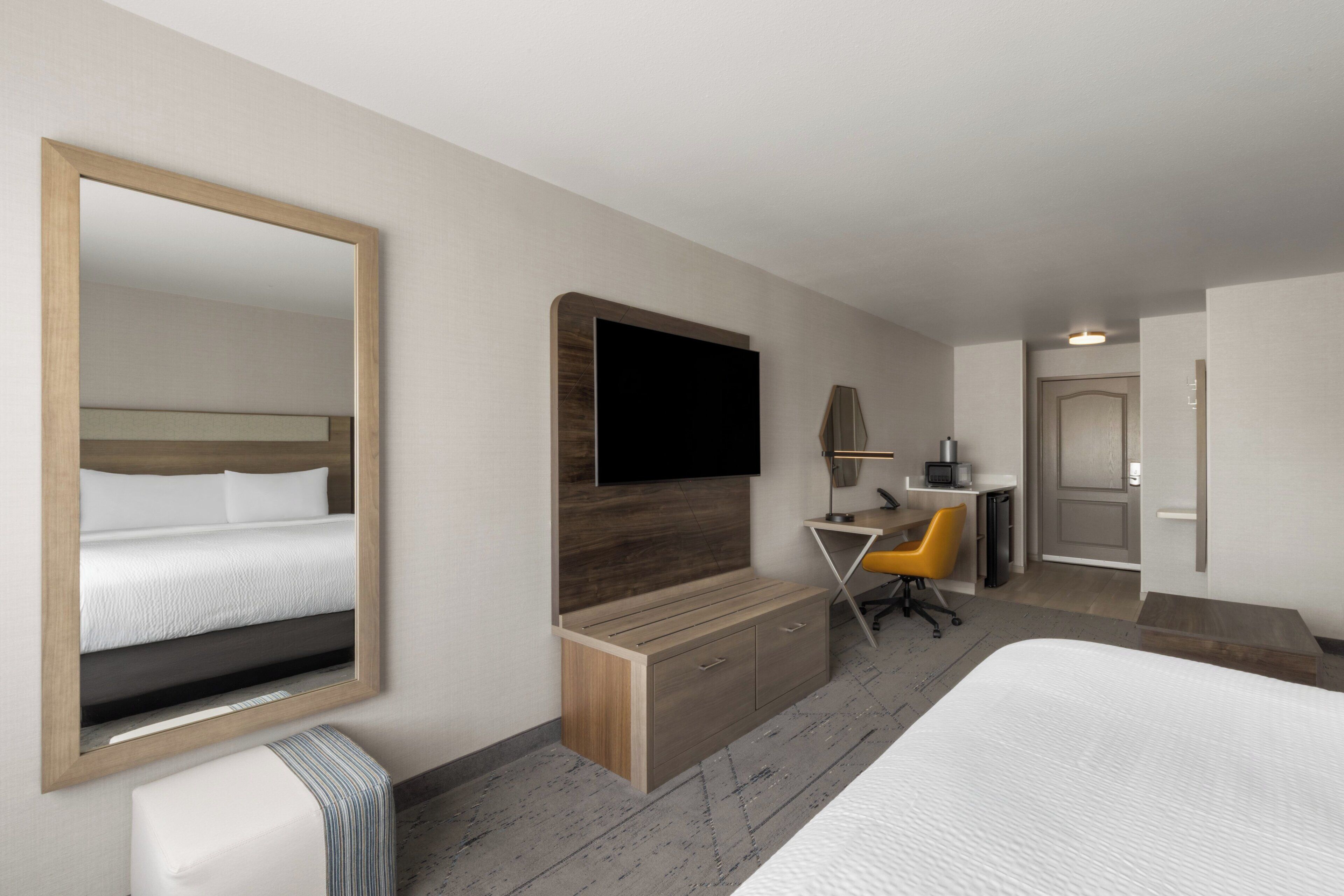 A hotel room with a large bed, a desk with a chair, a flat-screen TV mounted on the wall, and a mirror.