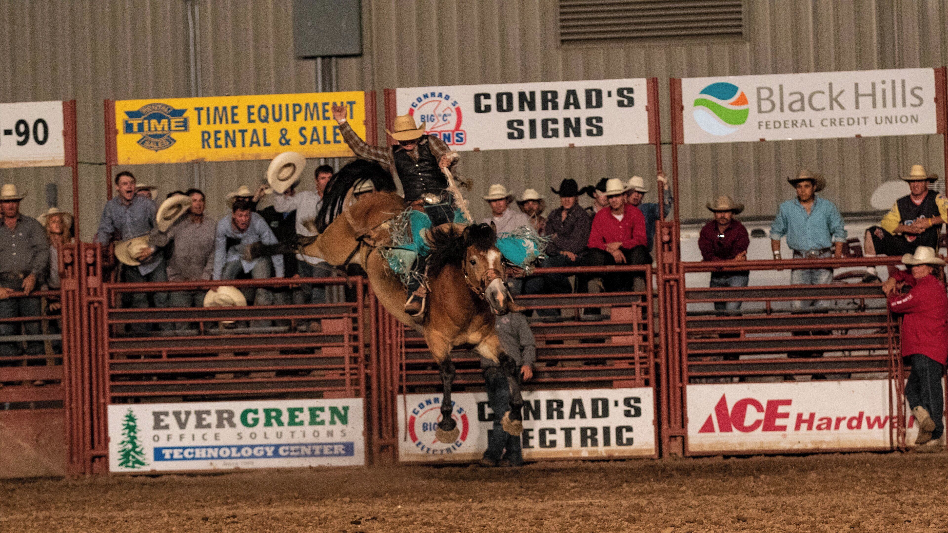 A rodeo event with a rider on a bucking horse, surrounded by spectators and various signage.