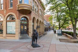 Une statue se trouve sur un trottoir, devant un immeuble doté de fenêtres en arc de cercle et d’une façade en briques.