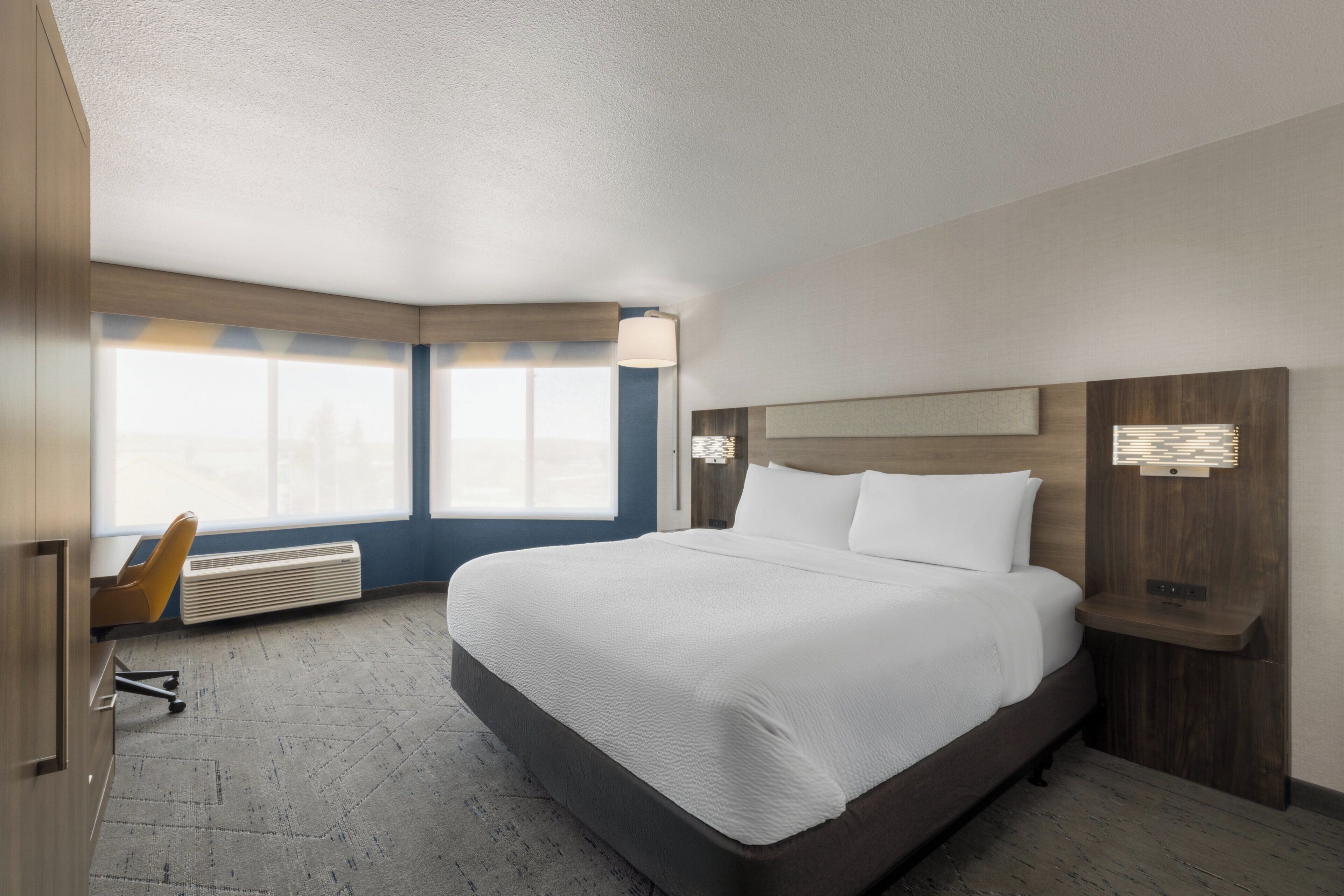 Una habitación de hotel con una cama grande, un escritorio con silla, una ventana con persianas y una lámpara fijada a la pared.
