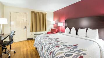 Ein Hotelzimmer mit Bett, Schreibtisch, Stuhl, einem Fenster mit Vorhängen und einer Wand mit rotem Akzent.