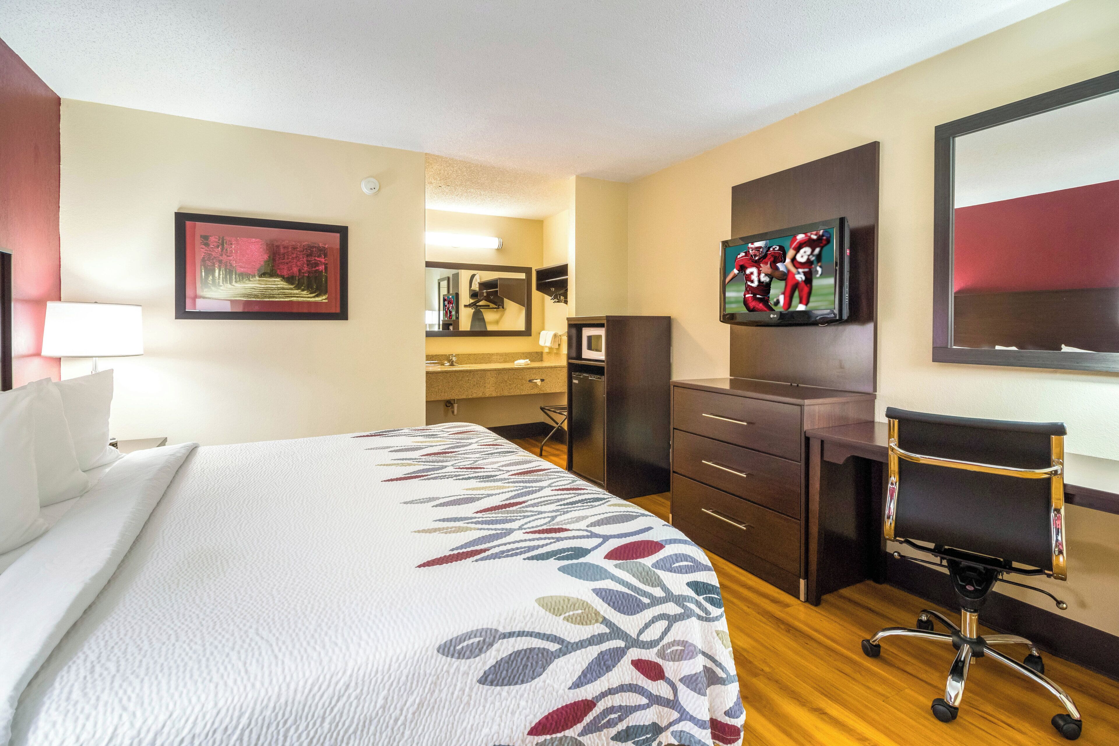 A hotel room with a large bed, a desk, a chair, a television, and a bathroom visible in the background.