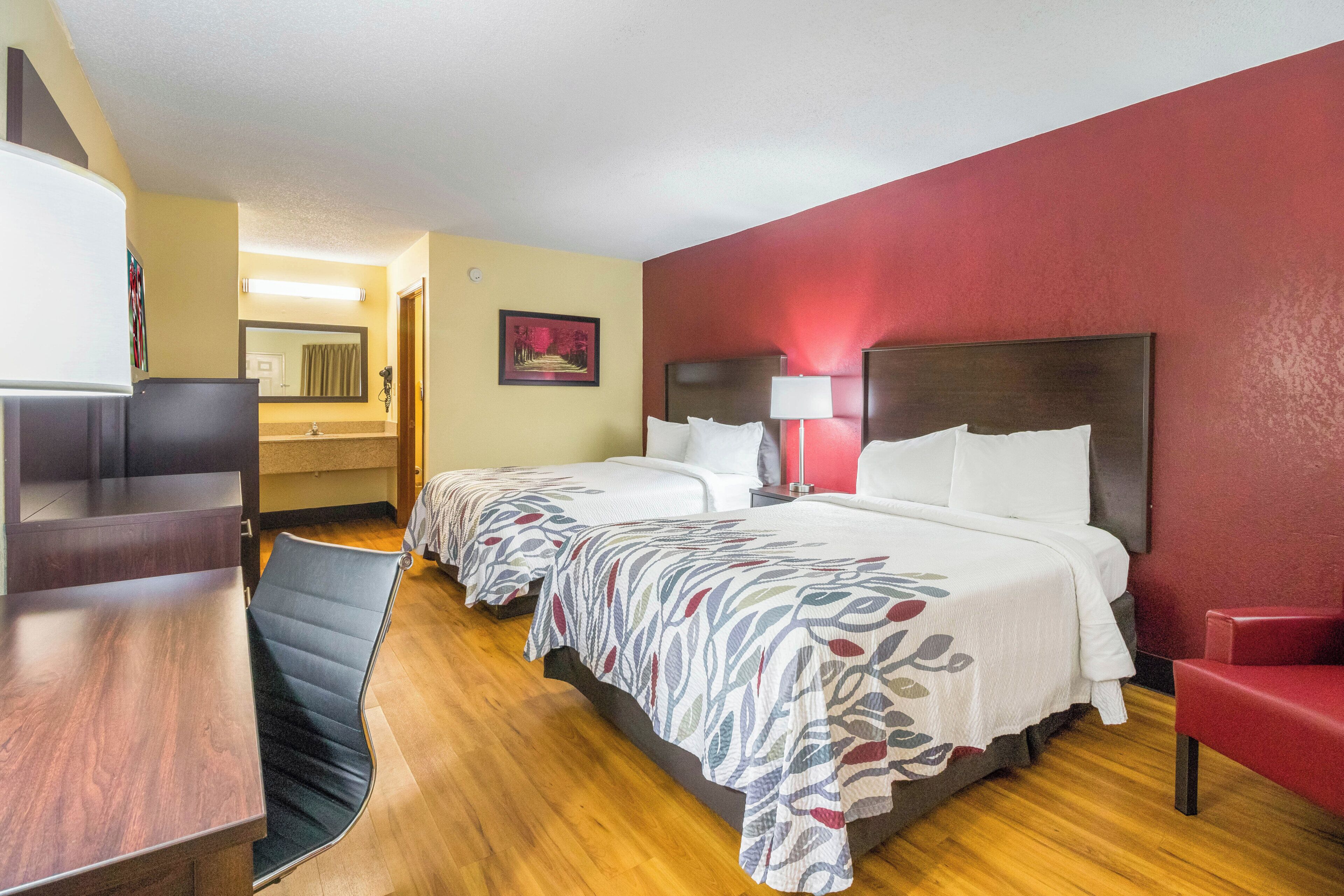 A hotel room with two beds, a desk, a chair, a mirror, a painting, and a red wall.