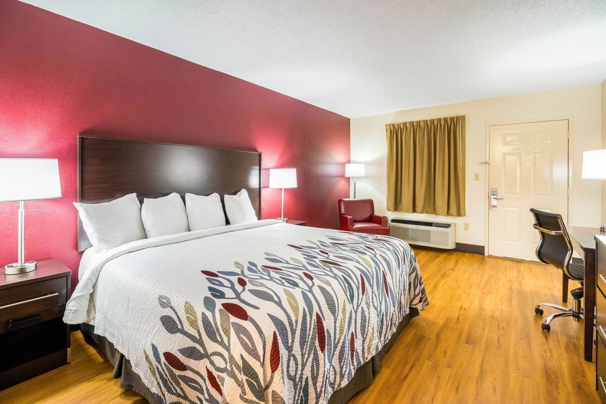 A hotel room with a bed, bedside tables, a desk, a chair, a red wall, and a door.