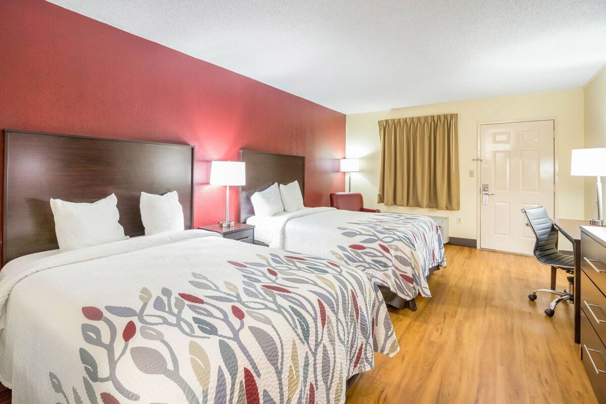 A hotel room with two beds, a desk, a chair, and a door.