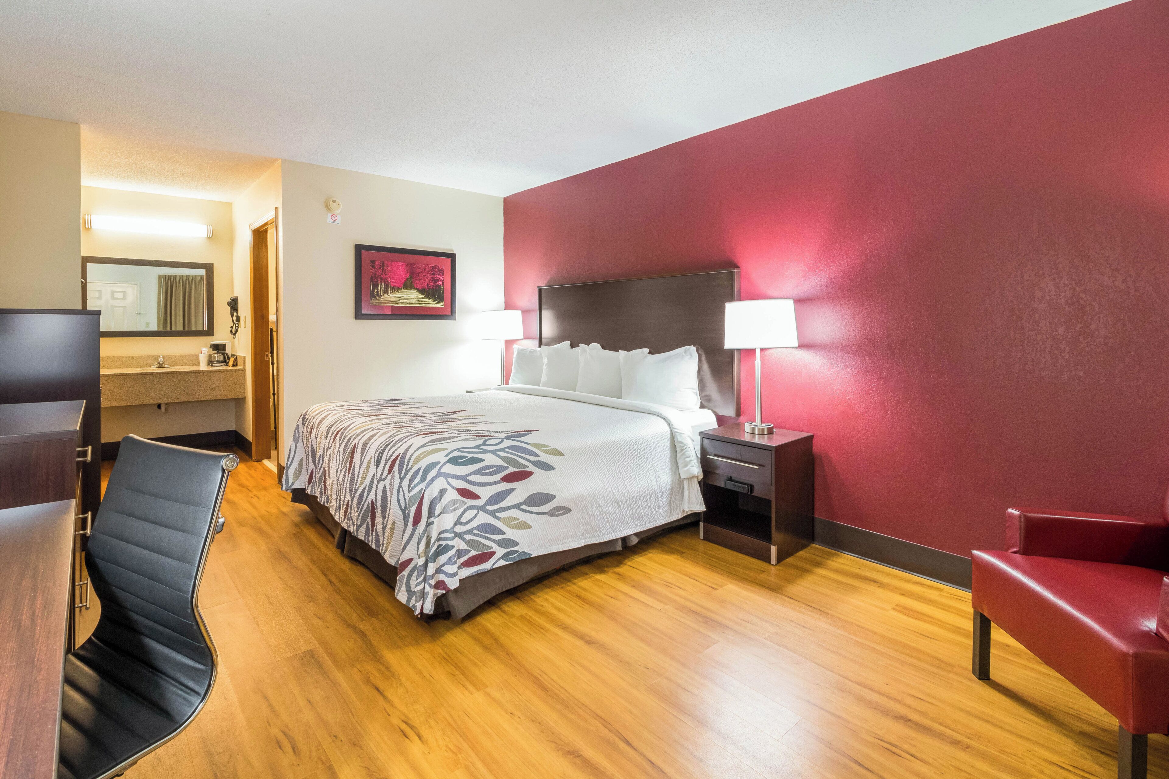 A hotel room with a bed, a desk, a chair, a nightstand, and a lamp.
