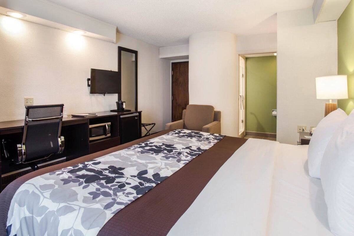 A hotel room with a bed, a desk with a computer, a chair, a television, and a door leading to another room.