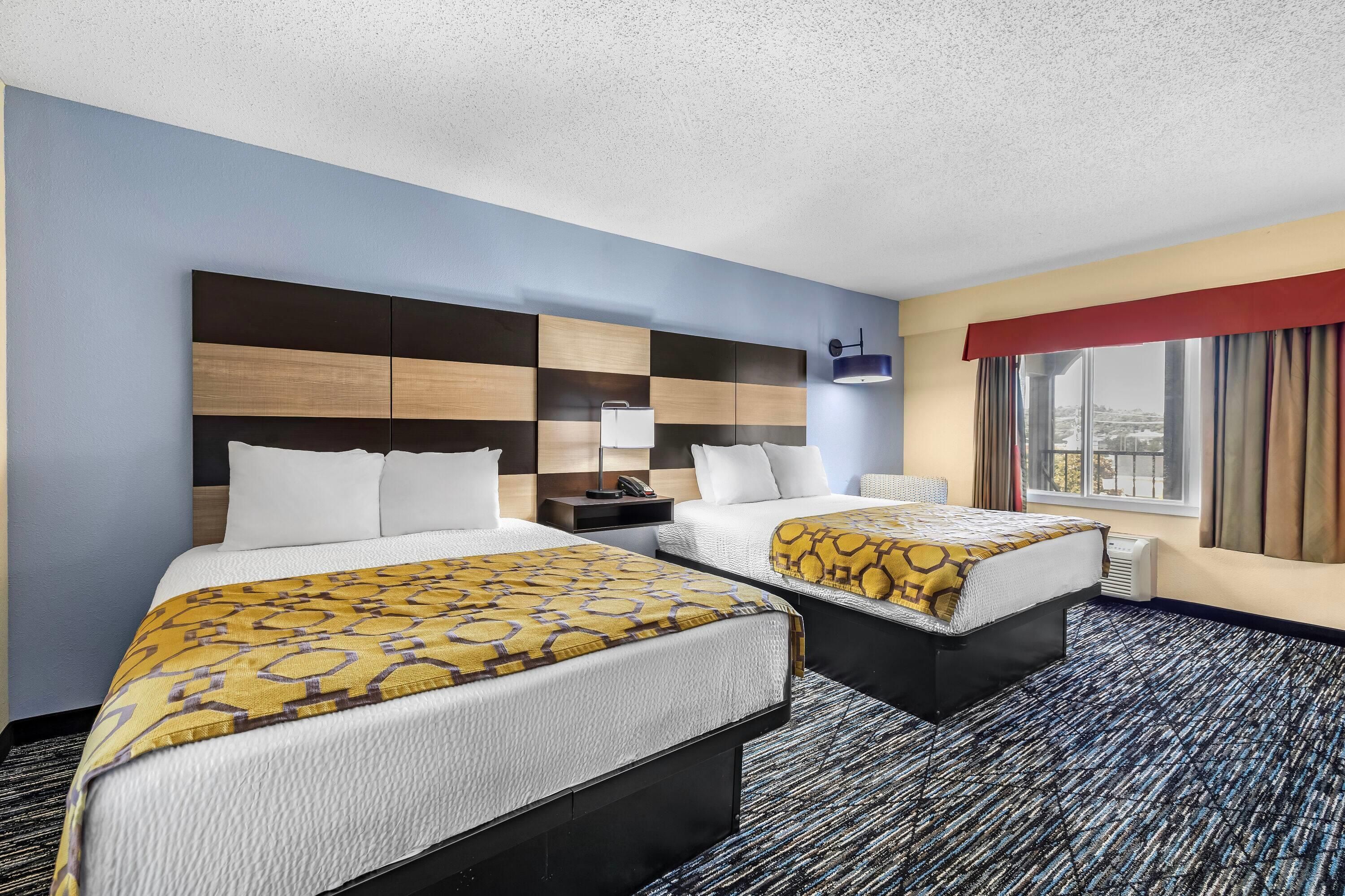 A hotel room with two beds, a headboard with a checkered pattern, a bedside table with a lamp, and a window with curtains.