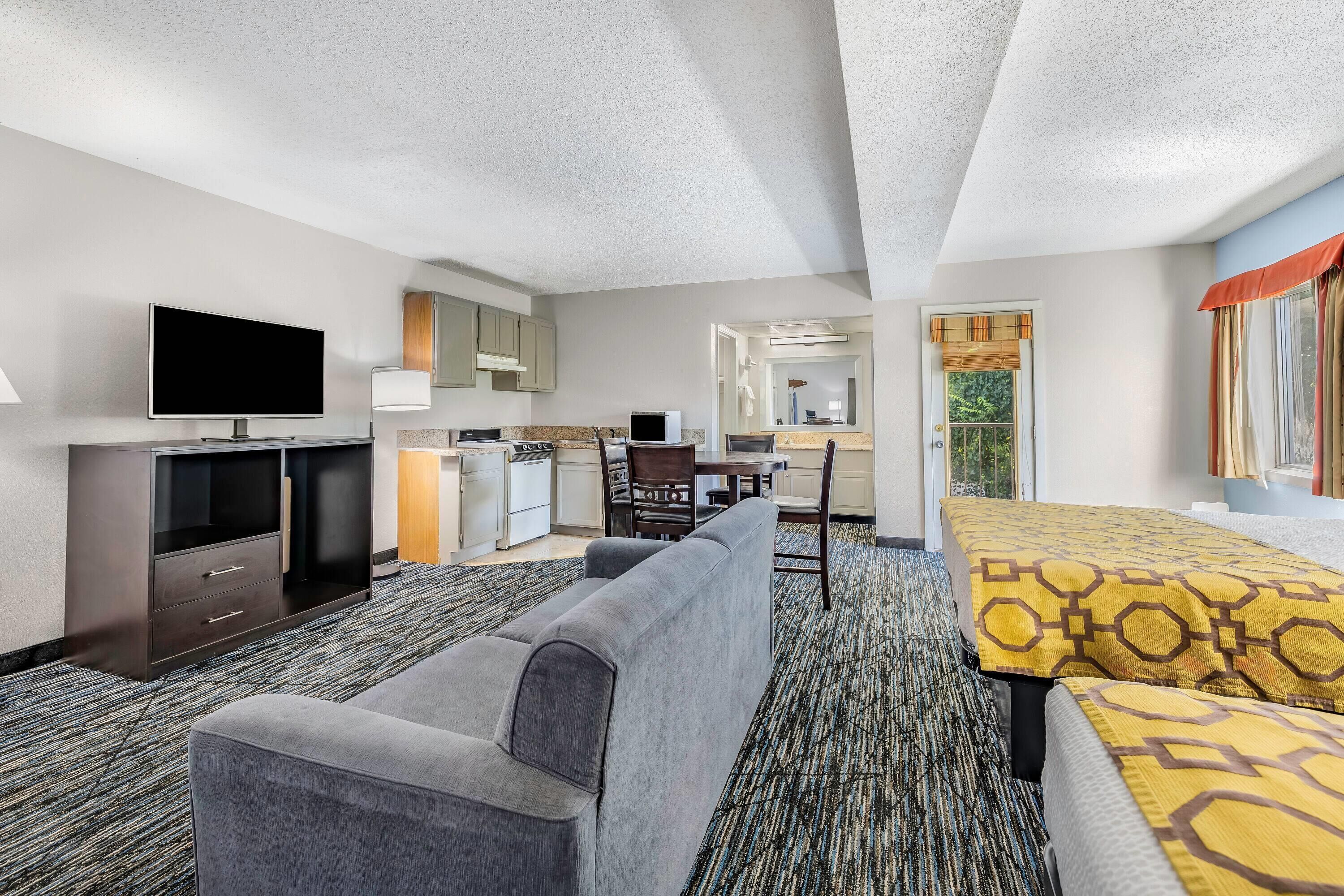 A hotel room with a flat-screen TV, a sofa, a bed with patterned bedding, a dining area with a table and chairs, and a kitchen area.