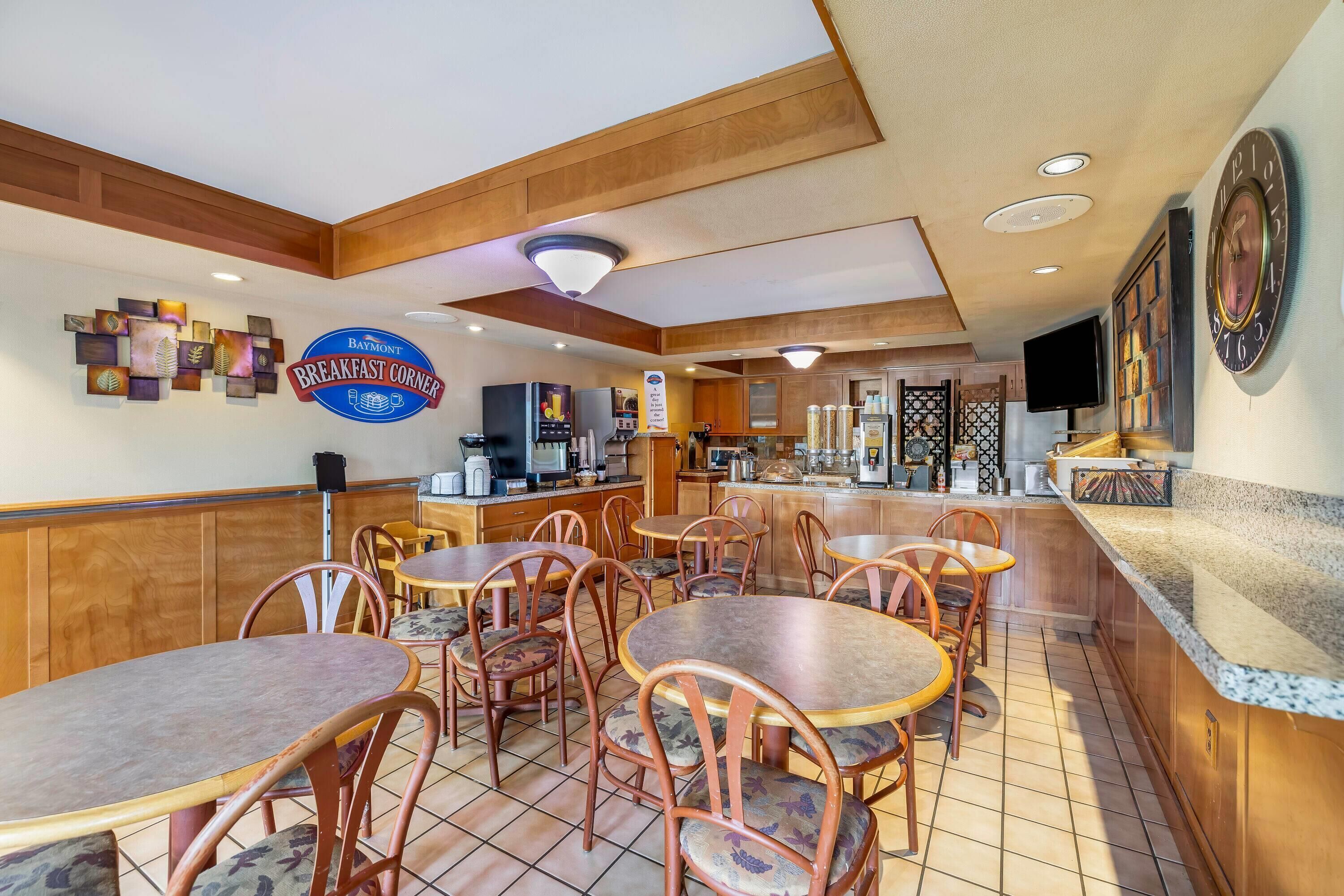A breakfast area with wooden tables and chairs, a Baymont Breakfast Corner sign, a coffee machine, and a television mounted on the wall.