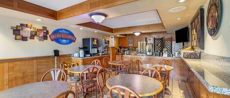 A breakfast area with wooden tables and chairs, a Baymont Breakfast Corner sign, a coffee machine, and a television mounted on the wall.