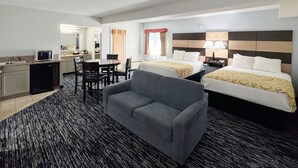 Une chambre d’hôtel avec une kitchenette, deux lits, un canapé gris et une grande fenêtre.
