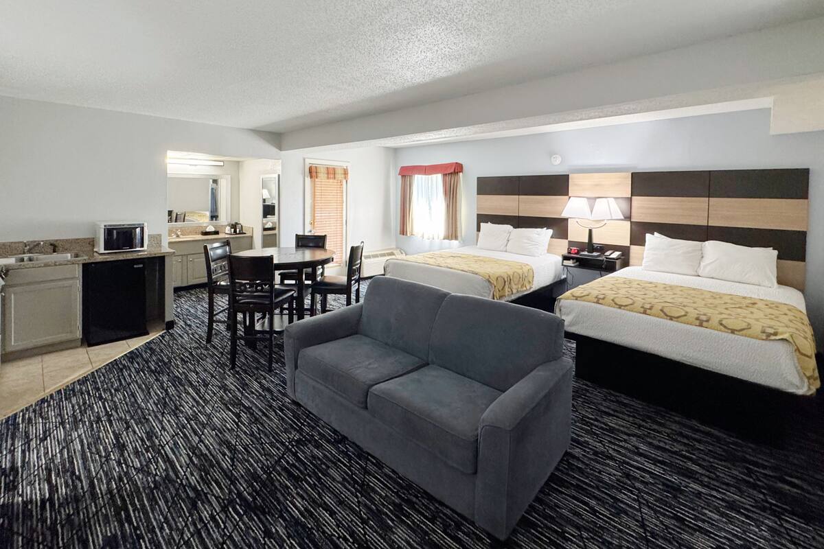 Een hotelkamer met kitchenette, twee bedden, een grijze bank en een groot raam.