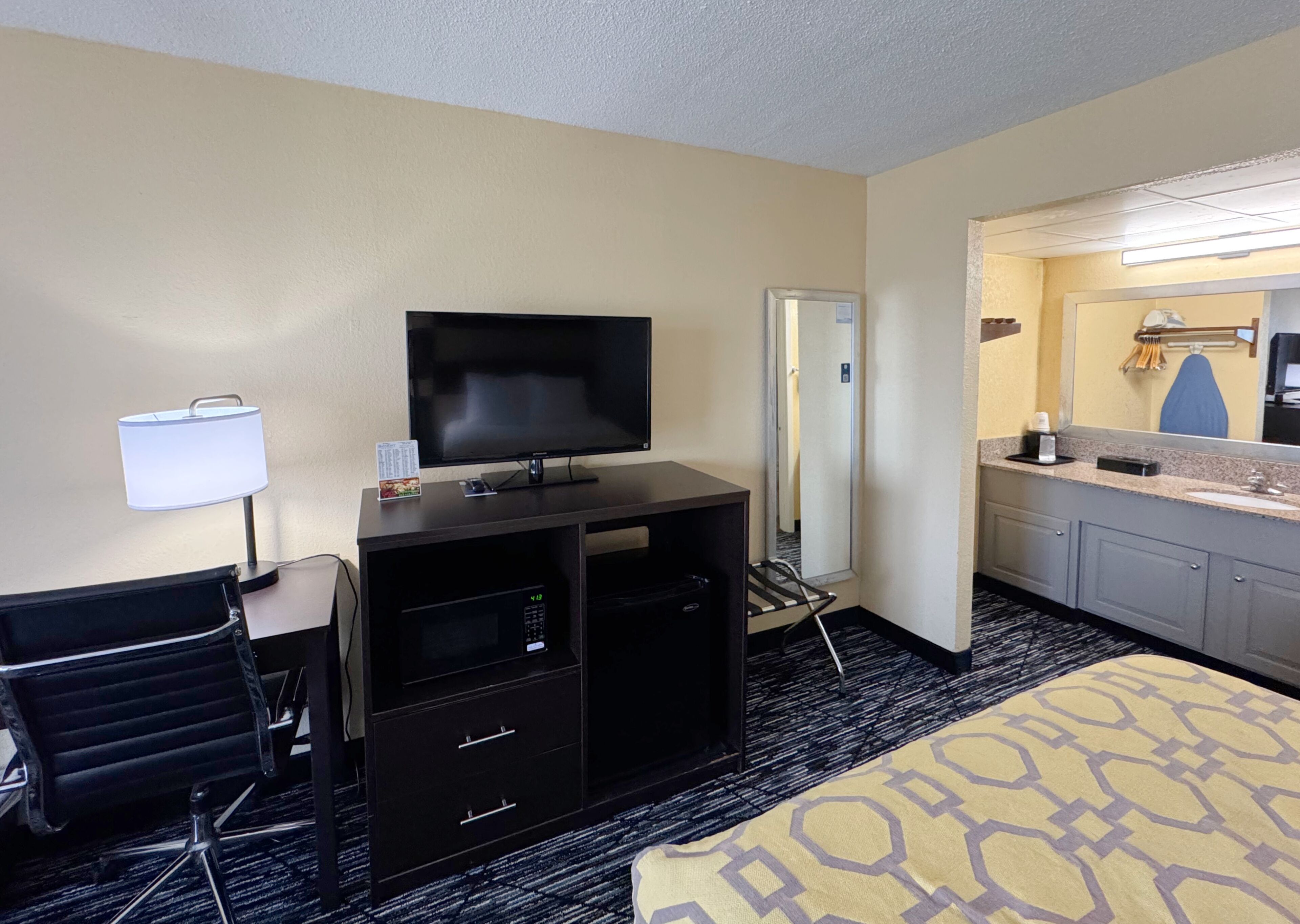 Camera d'albergo con televisione, scrivania, sedia, minibar e bagno con lavandino e specchio.